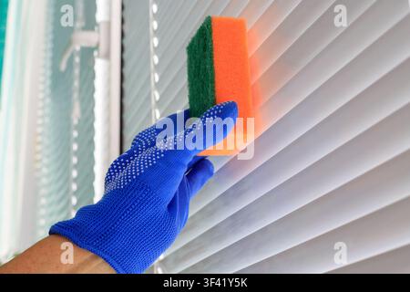 Cleaning blinds by hand. Person wipes white plastic slats with an abrasive orange-green sponge in an apartment in daylight. Stock Photo