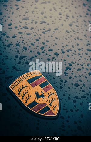 Close-up of the iconic gold Porsche crest badge on the front hood of a black sports car covered in raindrops Stock Photo