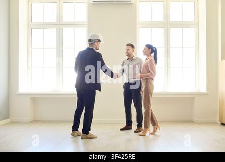 Couple discussing real estate with contractor, handshake in empty bright apartment Stock Photo