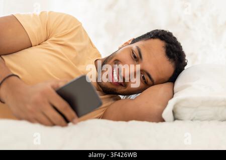 Smiling Young Black Man Using Smartphone While Lying On Bed, Relaxed Indoor Setting Stock Photo