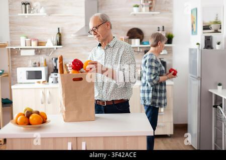 Senior couple arrives from the supermarket and unpacks grocery bags in the kitchen, helping each other during a relaxed morning routine. Stock Photo