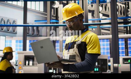 Professional working in smart factory, testing automated systems to ensure reliability. African american man in industry 4.0 plant using notebook to identify bottlenecks and optimize, camera A Stock Photo