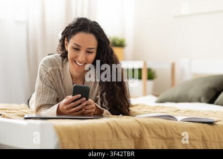 Happy Young Woman Using Smartphone Lying On Bed At Home, Relaxation Concept Stock Photo