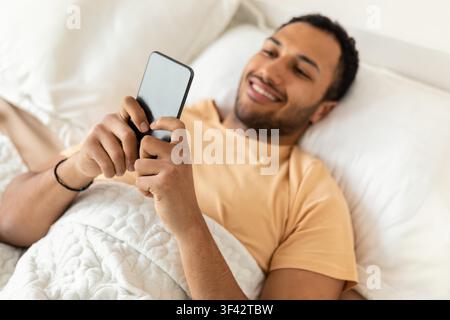Smiling Young African American Man Using Smartphone While Lying In Bed, Relaxing At Home Stock Photo