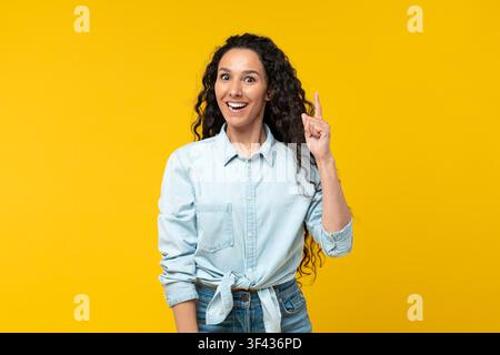 Smiling Young Hispanic Woman With Curly Hair Pointing Up At Yellow Background, Business Concept Stock Photo
