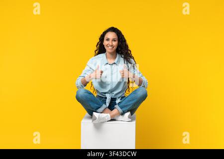 Happy Young Hispanic Woman Sitting On Box Giving Thumbs Up, Joyful Expression, Lifestyle Concept Stock Photo