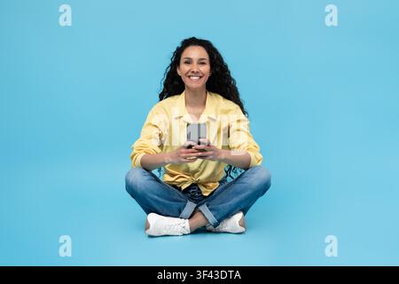 Happy Young Woman Sitting Cross Legged On Floor With Smartphone, Blue Background Stock Photo