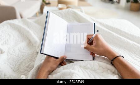 Unrecognizable Person Writing In Notebook While Sitting Comfortable On Bed At Home Stock Photo