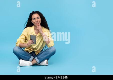 Thoughtful Young Latina Woman Using Smartphone While Sitting On Floor, Lifestyle Concept Stock Photo