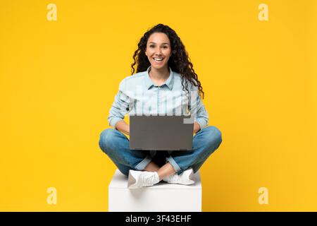 Smiling Young Mixed Race Woman Working On Laptop, Sitting On Cube, Yellow Background Stock Photo