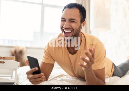 Happy Young Man Enjoying Phone Conversation, Laying On Bed, White Indoor Setting Stock Photo