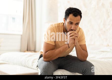 Worried Young Black Man Sitting On Bed In Bedroom, Deep In Thought, Mental Health Concept Stock Photo