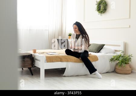 Happy Young Woman Working On Laptop Sitting On Bed At Home, Remote Work Concept Stock Photo