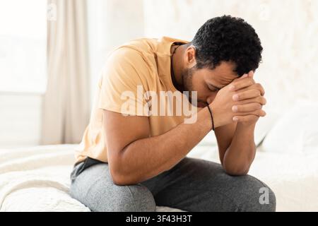 Worried Young Black Man Sitting On Bed With Hands Clasped, Mental Health Concept Stock Photo