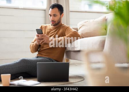 Young Man Sitting On Floor Using Smartphone, Working From Home, Comfortable Setting Stock Photo