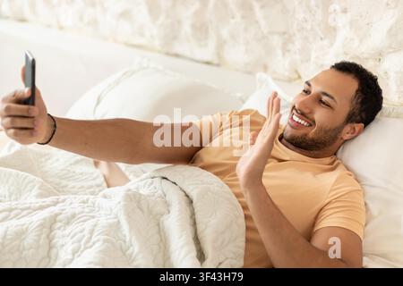 Happy Black Man Video Calling From Bed With Smartphone, Smiling And Relaxed, Home Lifestyle Concept Stock Photo