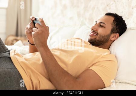 Smiling Young Black Man Relaxing On Bed While Playing Video Games, Home Lifestyle Stock Photo