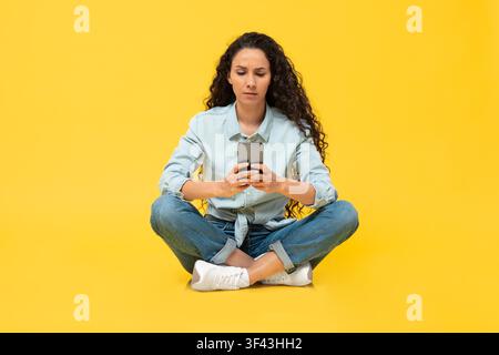 Focused Hispanic Woman Sitting Cross-legged Using Smartphone, Yellow Background Stock Photo