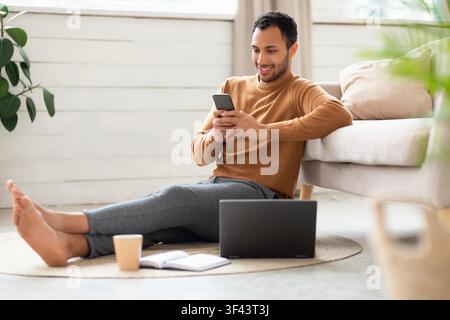 Smiling Young Hispanic Man Using Smartphone While Sitting On Floor In Cozy Living Room Stock Photo