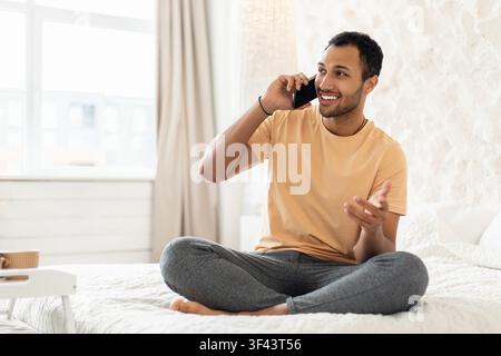 Smiling African American Man Sitting On Bed Talking On Phone, Home Lifestyle Concept Stock Photo