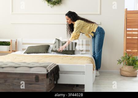 Woman In Yellow Sweater Arranging Green Pillows On Bed In Modern Bedroom, Cozy Home Concept Stock Photo