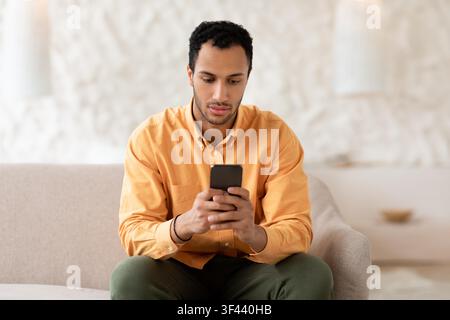 Focused Young Black Man Using Smartphone While Sitting On Couch, Indoor Setting Stock Photo