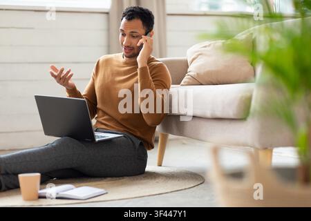 Focused Young Black Man Talking On Smartphone While Using Laptop At Home, Business Concept Stock Photo