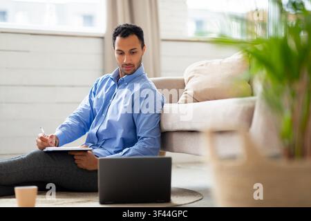 Focused Hispanic Male Student Taking Notes While Using Laptop On Floor At Home, Study Concept Stock Photo