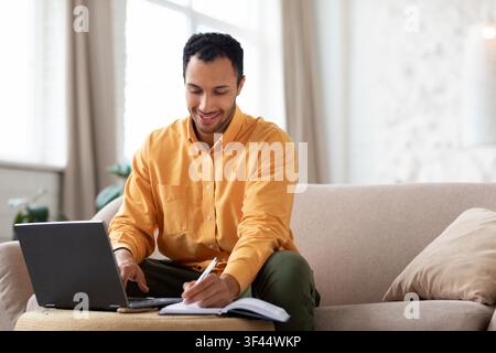Smiling Black Man Working On Laptop And Taking Notes At Home, Business Concept Stock Photo