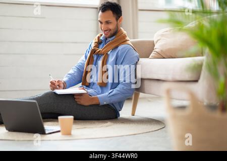 Smiling Young Man Writing Notes While Sitting On Floor With Laptop, Home Office Concept Stock Photo