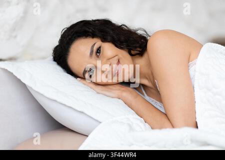 Smiling Young Middle Eastern Woman Relaxing On Bed With Pillow, Cozy Indoor Setting Stock Photo