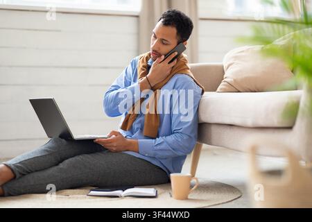Focused Black Man Working On Laptop While Talking On Phone At Home, Business Concept Stock Photo