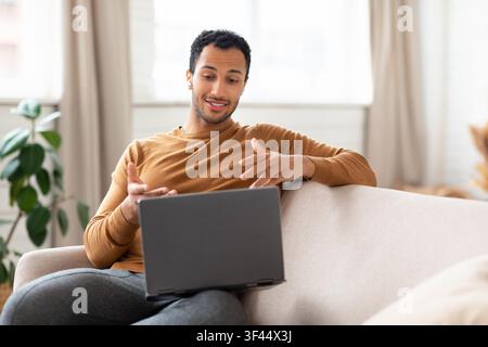 Happy Young Black Man Working On Laptop At Home, Engaged In Conversation, Business Concept Stock Photo