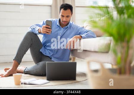 Focused Young Mixed Race Man Using Smartphone While Working On Laptop At Home, Business Concept Stock Photo
