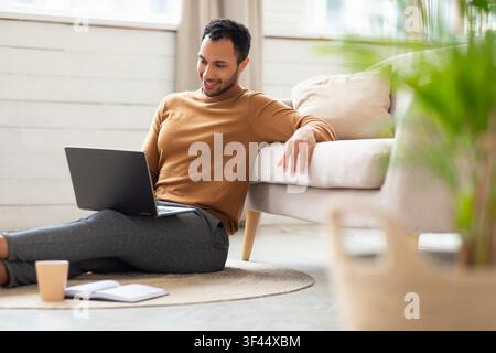 Smiling Young Black Man Working On Laptop While Sitting On Floor At Home, Professional Concept Stock Photo