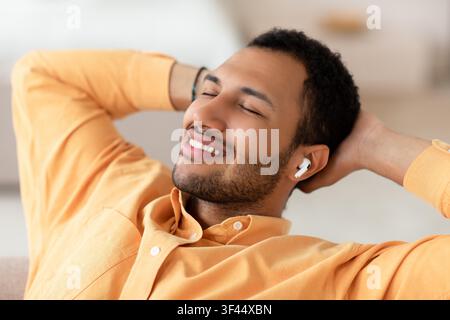 Smiling Young Black Man Relaxing With Wireless Earbuds At Home, Enjoying Music Stock Photo