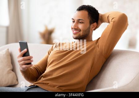 Smiling Young Man Relaxing On Sofa While Looking At Smartphone At Home Stock Photo