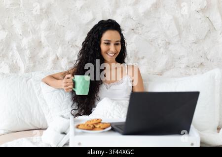 Smiling Young Hispanic Woman Enjoying Coffee And Cookies While Using Laptop At Home, Relaxation Concept Stock Photo