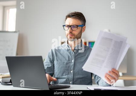 Focused Caucasian Male Working On Laptop, Holding Documents In Office, Business Concept Stock Photo