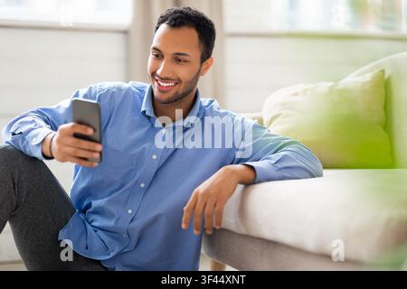 Smiling Young Man Using Smartphone While Relaxing At Home, Lifestyle Concept Stock Photo