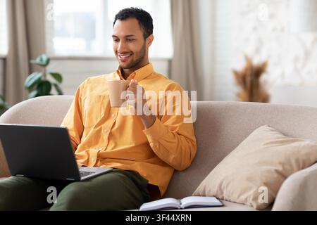 Smiling Young Man Using Laptop While Drinking Coffee At Home, Business Concept Stock Photo
