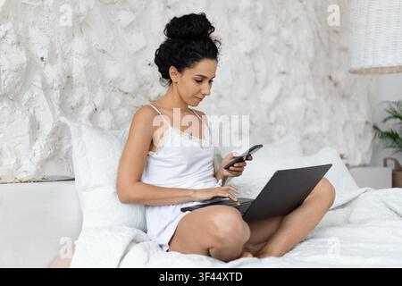 Focused Latin American Woman Using Laptop And Smartphone, Sitting On Bed, Home Office Concept Stock Photo