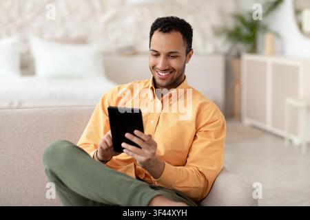 Smiling Young Black Man Using Tablet While Relaxing At Home, Lifestyle Concept Stock Photo