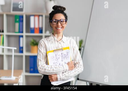 Smiling Young Latina Woman Holding Papers, Standing In Office, Business Concept Stock Photo