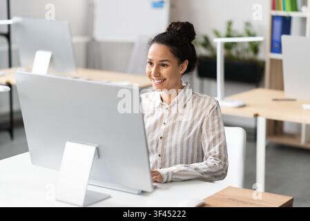 Smiling Young Hispanic Woman Working On Desktop Computer In Modern Office, Business Concept Stock Photo