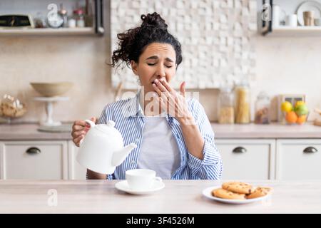 Tired Young Woman Yawning While Pouring Coffee In Kitchen, Morning Routine Stock Photo