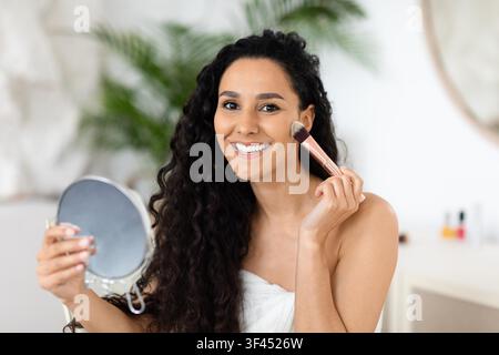 Smiling Young Woman Applying Makeup In Front Of Mirror At Home, Skincare Concept Stock Photo