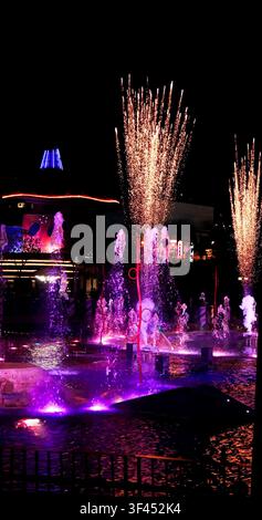 Dynamic water fountains illuminated in vibrant purple, pink, and orange, with bright sparkler effects against a dark night sky. Festive urban entertai Stock Photo