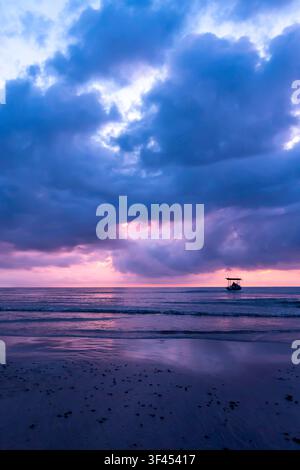 A lone boat sails across a tranquil ocean under a dramatic sky painted with vibrant purple, blue, and pink hues during a breathtaking sunrise. Reflect Stock Photo