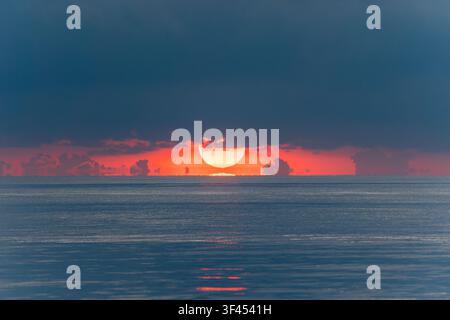 A breathtaking and dramatic ocean sunrise with a vibrant, fiery orange and red sky, showcasing the sun half-hidden behind dark clouds, reflecting soft Stock Photo